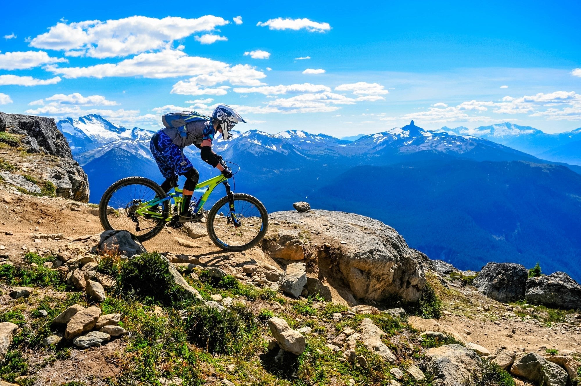 Mountain biker riding on a mountain with blue sky and mountains in background.