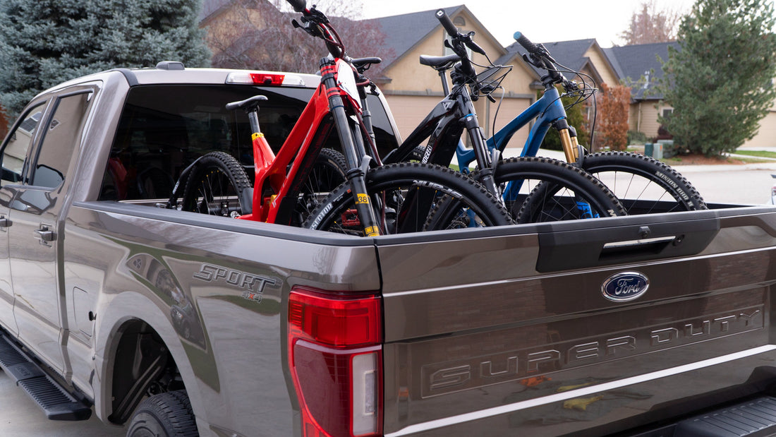 A grey Ford Super Duty truck with mountain bikes in RIDE88 QR3 Truck Bed Bike Racks. Suburban neighborhood with trees and houses in the background.
