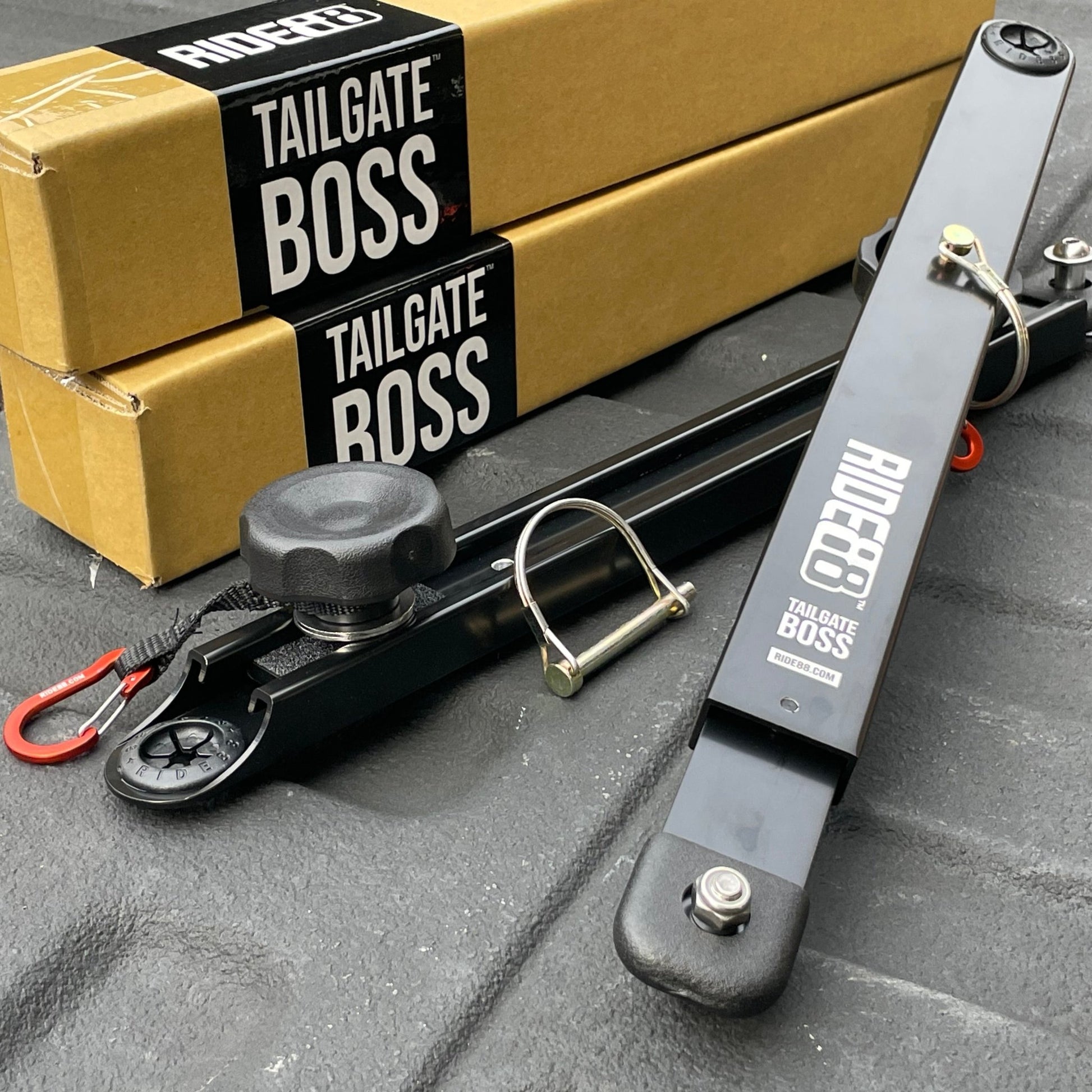 RIDE88 Tailgate Boss Pro-Kit tailgate support bars resting in front of cardboard boxes with black stickers with white text that says "RIDE88 TAILGATE BOSS".